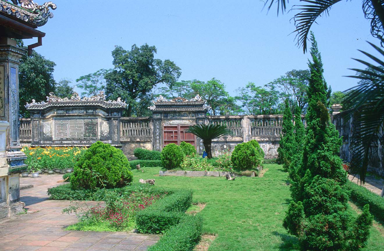 Ornate garden courtyard with manicured shrubs, palm trees, and traditional decorated stone walls and gate under blue sky